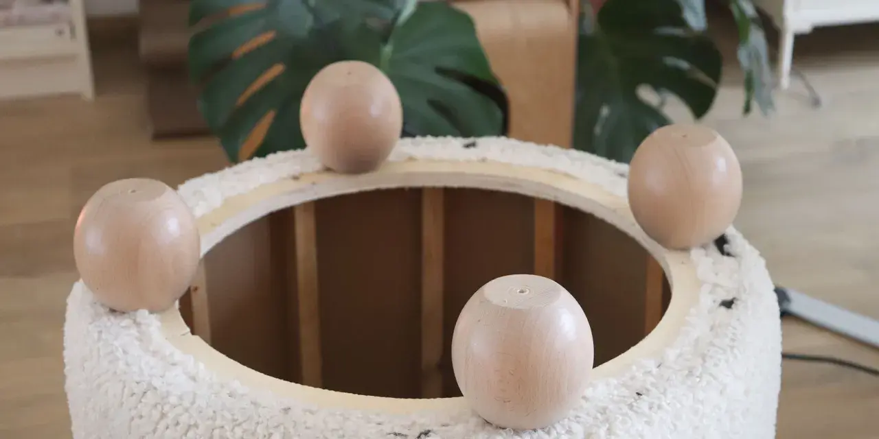 Quatre pieds ronds en bois sont posés sur le bord d'un pouf rond en tissu bouclette, prêt à recouvrir.