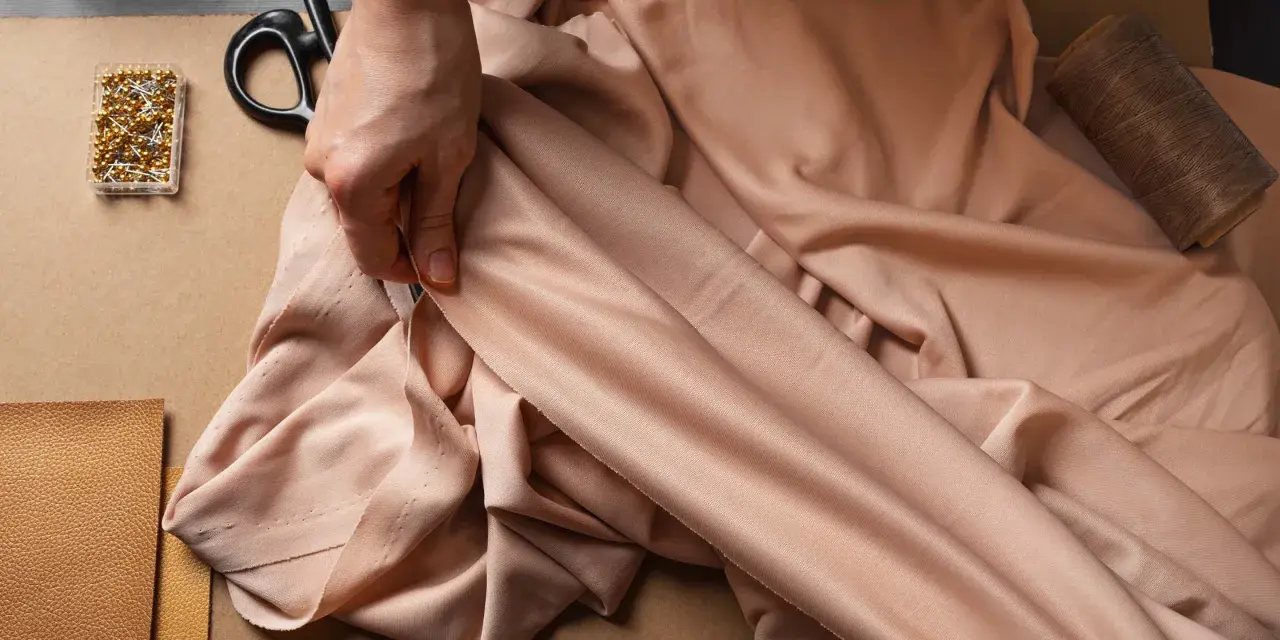 Mains tenant un tissu drapé, avec des épingles, des ciseaux et du fil. La viscose, c'est quoi ? Une matière douce et fluide pour la couture.