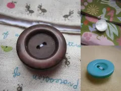 Gros plan sur un bouton à queue marron, prêt à être cousu sur un tissu orné de petites fourmis. Un autre bouton nacré est visible sur un tissu coloré.