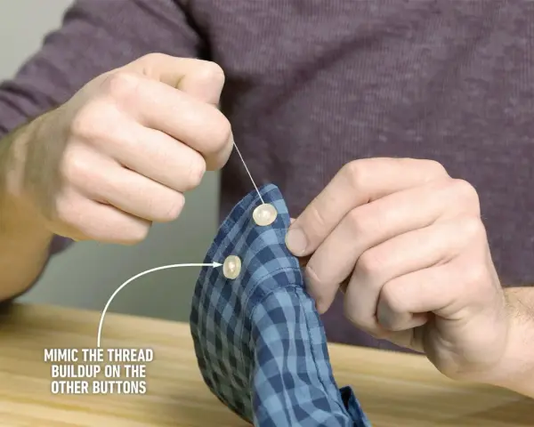 Main tenant une chemise à carreaux bleus, il est en train de coudre un bouton avec une aiguille et du fil blanc.