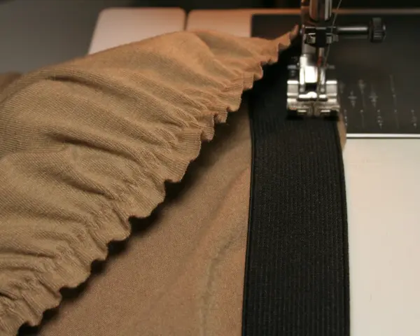 Machine à coudre en train de coudre un élastique sur un tissu beige froncé. Le pied presseur est visible, ainsi que l'aiguille.