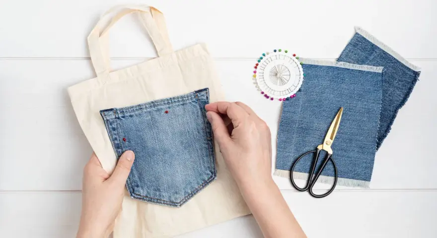 Mains épinglant une poche extérieure en jean sur un sac en toile. Ciseaux et chutes de tissu à proximité.