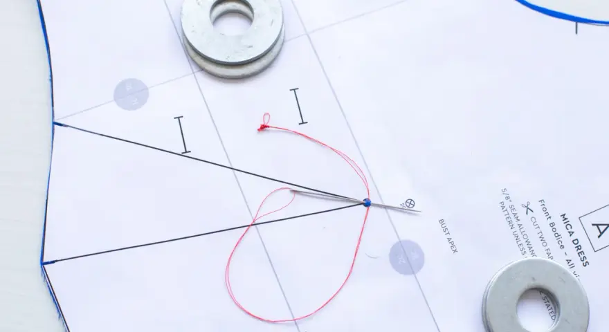 Sur un patron de couture, une aiguille rouge traverse le papier, retenue par des rondelles métalliques.