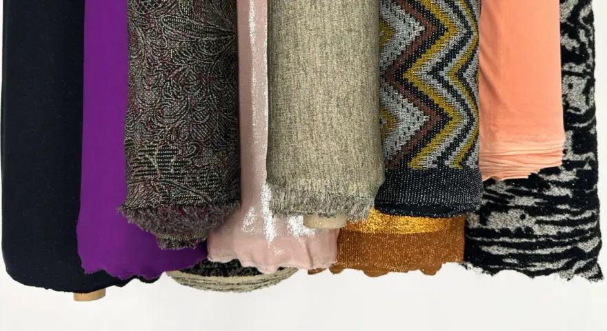 Rouleaux de tissus aux textures variées : noir uni, violet, motifs complexes, rose pâle, beige chiné, zigzags multicolores, pêche, et noir et blanc. Une matière mesh se distingue par sa finesse.