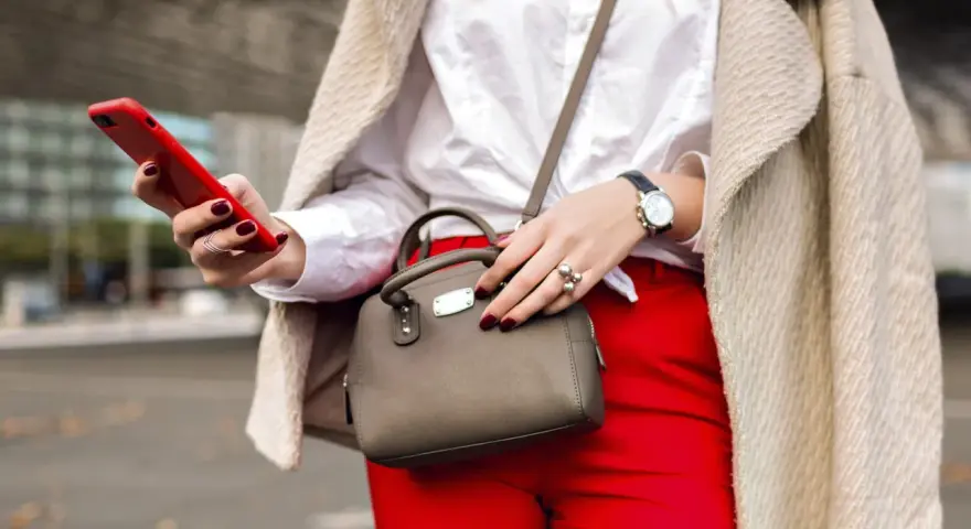 Une femme élégante, avec un manteau beige, un chemisier blanc et un pantalon rouge vif, tient un téléphone rouge et un petit sac gris. Quel entoilage pour sac choisir pour un tel look ?