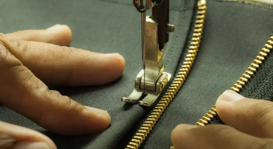 Main tenant une fermeture éclair dorée, une aiguille de machine à coudre s'apprête à coudre une fermeture éclair à la main sur un tissu sombre.