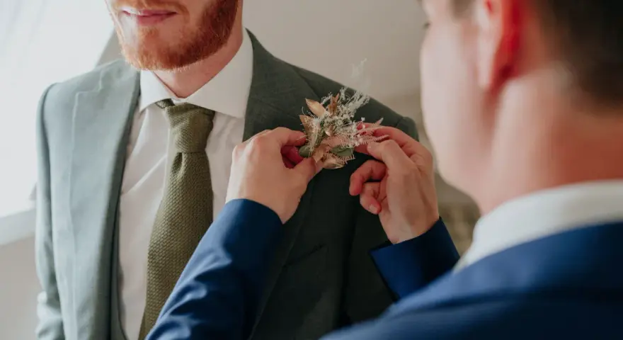 Un homme ajuste une boutonnière sur le costume d'un autre. Divers types de boutonnières existent, celle-ci est composée de fleurs séchées et de feuillages dorés.