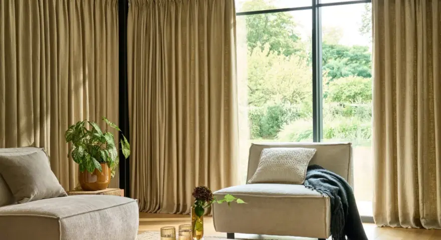 Salon moderne avec canapé beige, fauteuil, plante verte et rideaux au **type de pli rideau** qui habillent une grande baie vitrée.