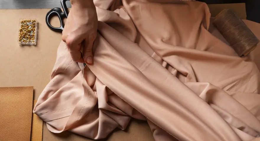 Mains tenant un tissu drapé, avec des épingles, des ciseaux et du fil. La viscose, c'est quoi ? Une matière douce et fluide pour la couture.