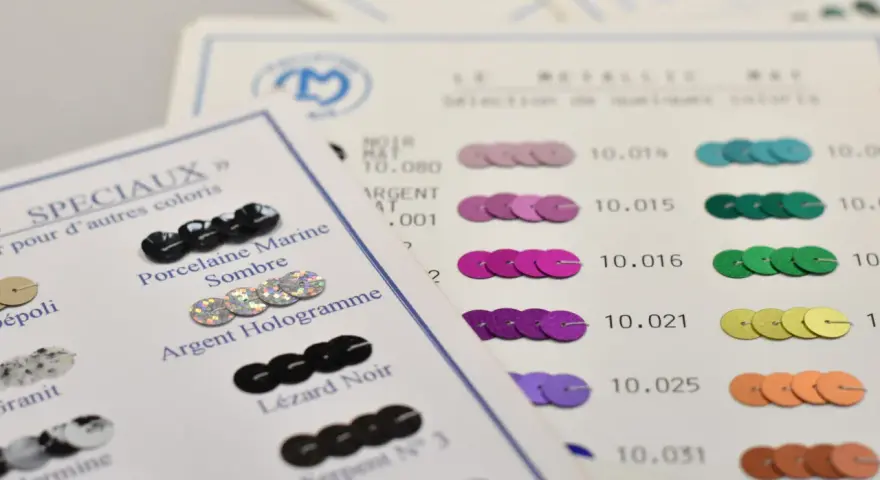 Échantillons de sequins pour faire un motif. Des sequins noirs, argentés, roses, violets, verts, jaunes et orangés sont présentés sur des fiches de couleurs.