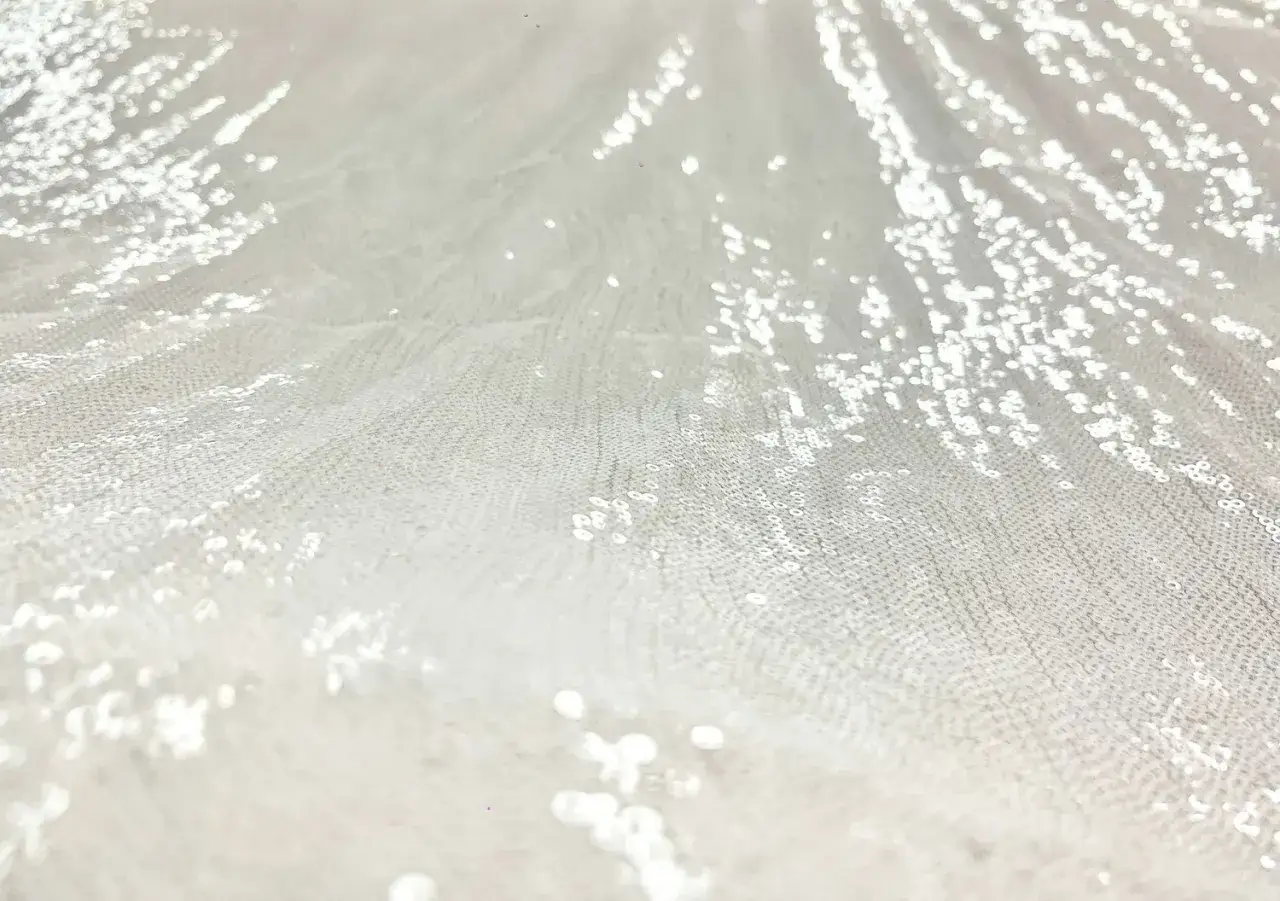 Tissu blanc avec un motif en sequin qui scintille, évoquant la neige ou des étoiles.