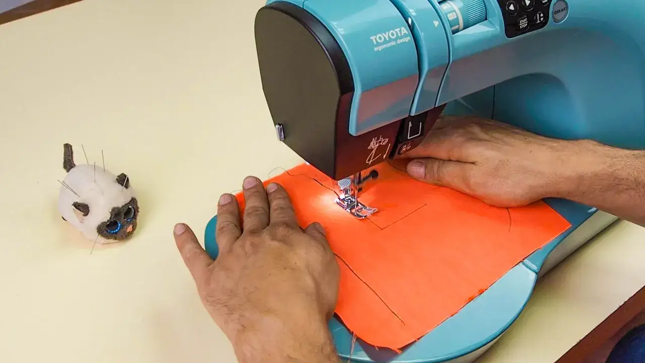 Apprendre à comment coudre avec une machine Toyota. Un petit chat en tissu, piqué d'aiguilles, observe la scène.